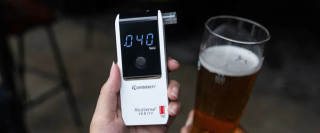 Hand holding AlcoSense Verity personal breathalyser with beer in background