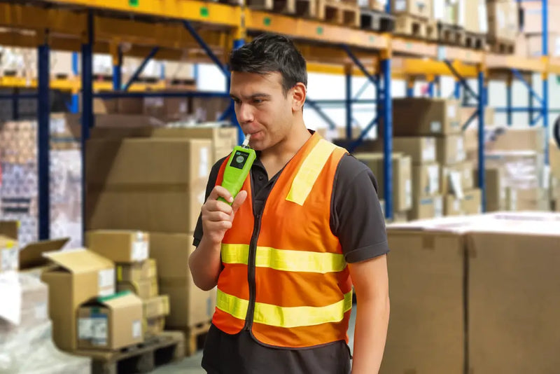 A warehouse worker wearing a safety vest stands in a storage facility, blowing into a handheld Andatech Vanguard professional breathalyser